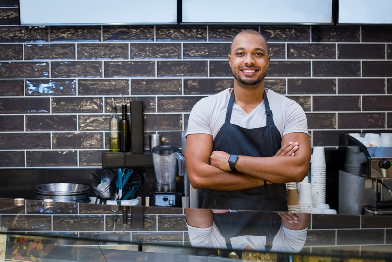 Portrait of a smiling waiter standing in the cafeteria with crossed arms. Confident young man with apron in coffee shop looking at camera. Handsome black guy working in a coffeeshop.